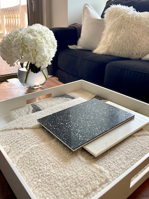 material samples in a tray on a table with a vase of flowers and a sofa nearby