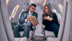 man pointing to tablet screen on business jet, woman looking at tablet screen with coffee cup in hands