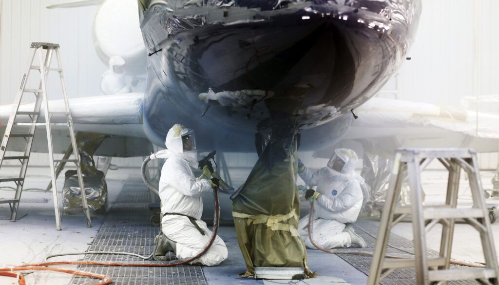 two people in protective clothing with paint equipment underneath nose of aircraft being painted