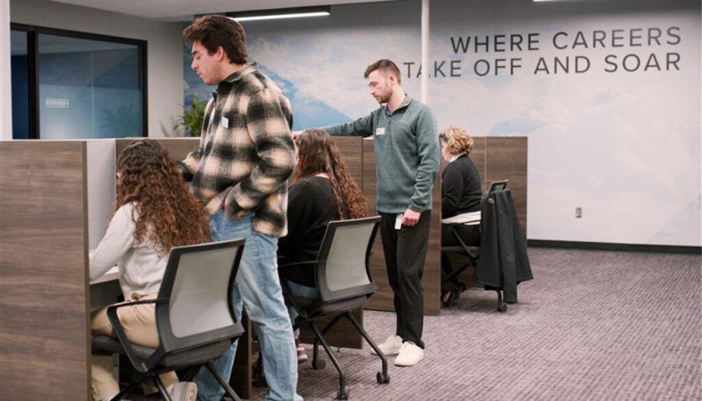 3 people seated at desks with dividers in-between, two people standing