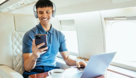 man sat on private jet using laptop, phone and headphones