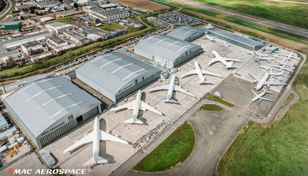 Aerial view of airport with lots of aircraft parked outside large buildings with grey roofs