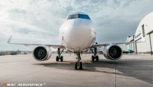 Front view of white aircraft on tarmac showing nose, engines, and landing gear outside hangar building