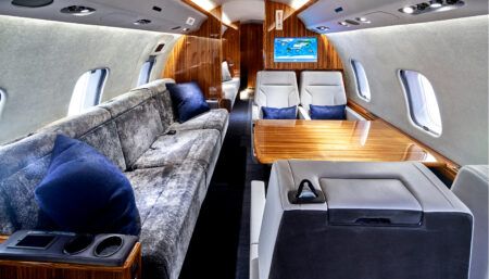 Private jet cabin with grey divan, white chairs, wood table, and entertainment screen visible