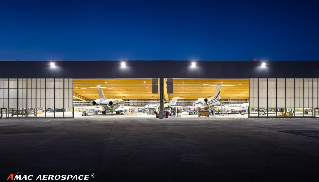 Night-time exterior view of illuminated aircraft hangar with open doors revealing multiple business jets inside under bright lighting