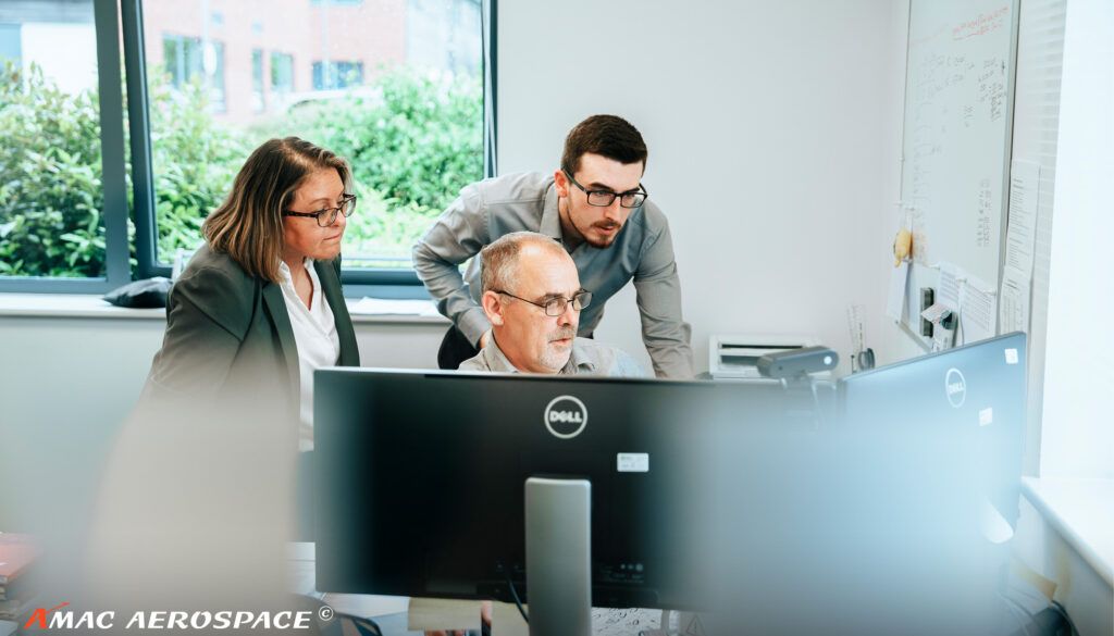Three business professionals collaborating around computer monitors in modern office with window view of greenery