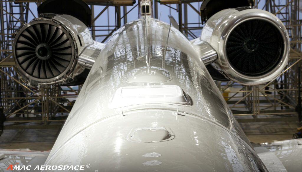 Close-up view of white aircraft fuselage in hangar, showing satcom equipment on roof, and twin jet engines