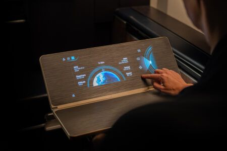 Hand touching tray table surface with integrated high-definition touchscreen display showing blue flight information graphics including moving map, departure and arrival airports, altitude, speed and time data