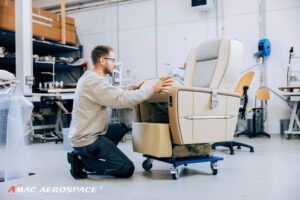 Technician in beige top working on business jet passenger seat with cream leather upholstery, grey insert and blue piping mounted on wheeled platform in aircraft completion workshop