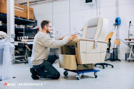 Interior modifications on head-of-state A340 project at AMAC Technician in beige top working on business jet passenger seat with cream leather upholstery, grey insert and blue piping mounted on wheeled platform in aircraft completion workshop