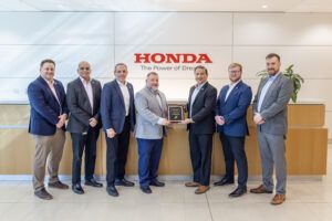 Seven business professionals standing in corporate reception area beneath Honda company logo and tagline, with two men in centre holding framed certificate