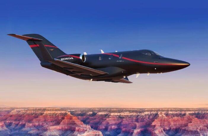HondaJet Elite II business jet in flight over canyon landscape at sunset, featuring black and red livery with Honda branding, and over-the-wing mounted engines