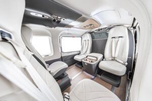 Wide-angle view inside an ACH145 helicopter cabin showing white curved cabin walls, quilted leather seats, wood flooring, metallic accents, central console with two cupholders and large windows on both sides.