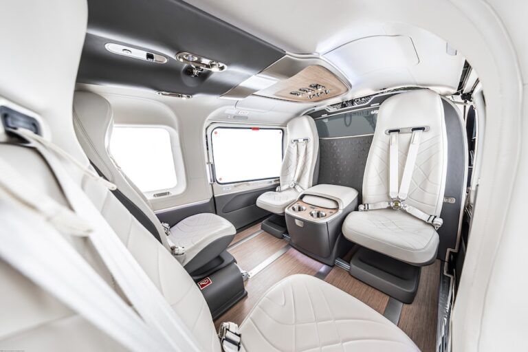 Wide-angle view inside an ACH145 helicopter cabin showing white curved cabin walls, quilted leather seats, wood flooring, metallic accents, central console with two cupholders and large windows on both sides.