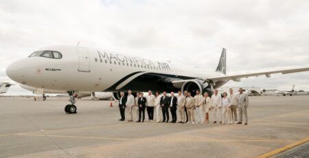 A group of 17 people stand on an airport apron in front of an ACJ321neo aircraft bearing the Magnifica Air name and livery, featuring a white base and black band on lower fuselage.
