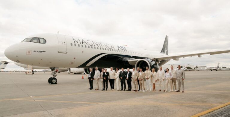 A group of 17 people stand on an airport apron in front of an ACJ321neo aircraft bearing the Magnifica Air name and livery, featuring a white base and black band on lower fuselage.