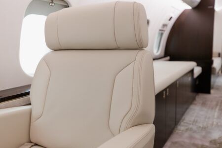 Close-up detail of beige business jet passenger seat showing perforated leather centre panel with smooth leather bolsters and headrest, aircraft window and dark wood sidewall visible in background