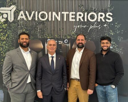 Four men stand in front of an Aviointeriors branded wall featuring the company logo and the tagline “your plus +”