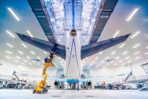 Wide-angle view from beneath the tail of a large aircraft in a brightly lit paint hangar, with technician on a scissor lift working on the fuselage.
