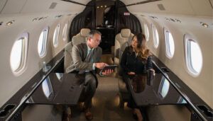 Two business professionals in suits seated opposite each other in beige leather club seats aboard business jet cabin, man holding tablet showing woman screen content, cream-coloured interior with dark wood accents