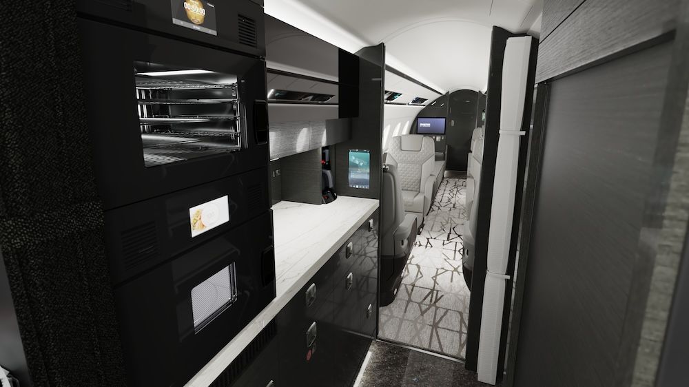 Praetor 600E business jet galley with black cabinetry, white worktop, ovens, and storage drawers, opening into cabin with patterned carpet.