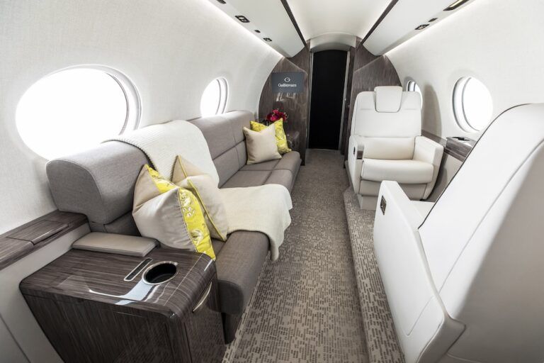 View looking aft along the Gulfstream G300 cabin showing a grey fabric divan with cream and yellow cushions and a cream knitted throw on the left, two white leather club seats on the right, dark wood-veneer trim and a cup holder console.