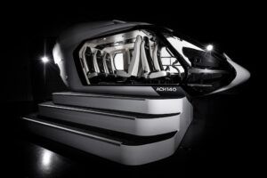 Photograph of the Airbus ACH140 helicopter cabin mock-up, open on one side, showing the full passenger compartment with white and dark grey seats. Steps leading up to the open side.