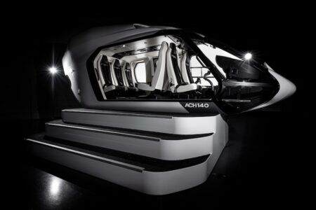 Photograph of the Airbus ACH140 helicopter cabin mock-up, open on one side, showing the full passenger compartment with white and dark grey seats. Steps leading up to the open side.