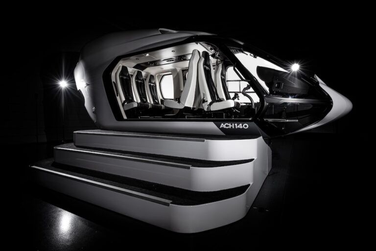 Photograph of the Airbus ACH140 helicopter cabin mock-up, open on one side, showing the full passenger compartment with white and dark grey seats. Steps leading up to the open side.