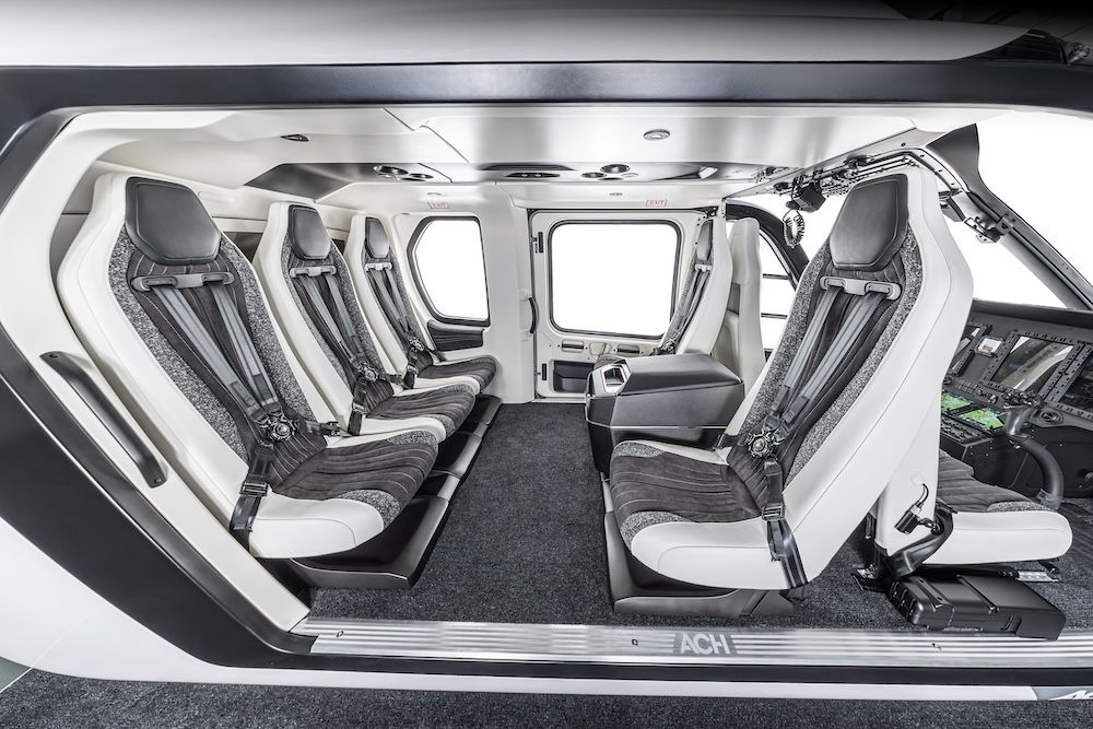 Interior view of the Airbus ACH140 helicopter cabin mock-up, showing five white and grey upholstered seats with four-point harnesses arranged facing each other, a cabinet, large windows and an open cockpit area to the right.