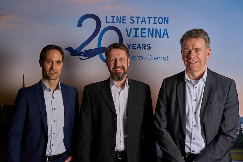 Three men stand smiling in front of a branded backdrop reading “Line Station Vienna 20 Years Aero-Dienst”.