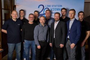 A group of 10 men stand together in front of a branded backdrop reading “Line Station Vienna 20 Years Aero-Dienst”.