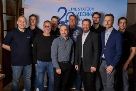 A group of 10 men stand together in front of a branded backdrop reading “Line Station Vienna 20 Years Aero-Dienst”.
