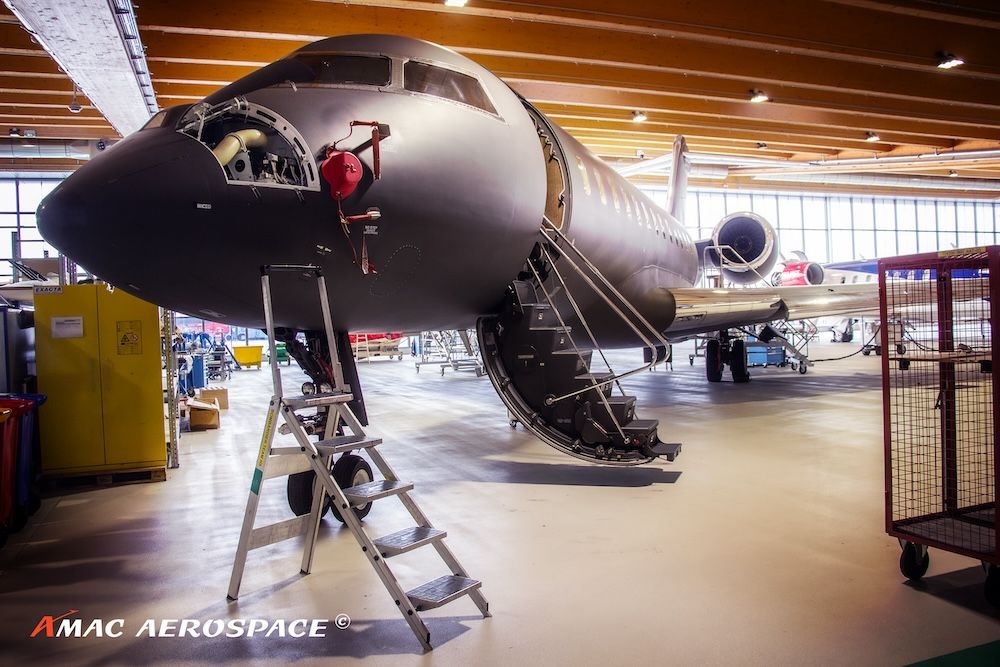 A Bombardier Global Express XRS aircraft inside a maintenance hangar, with an access ladder positioned at the nose and the cabin door open.