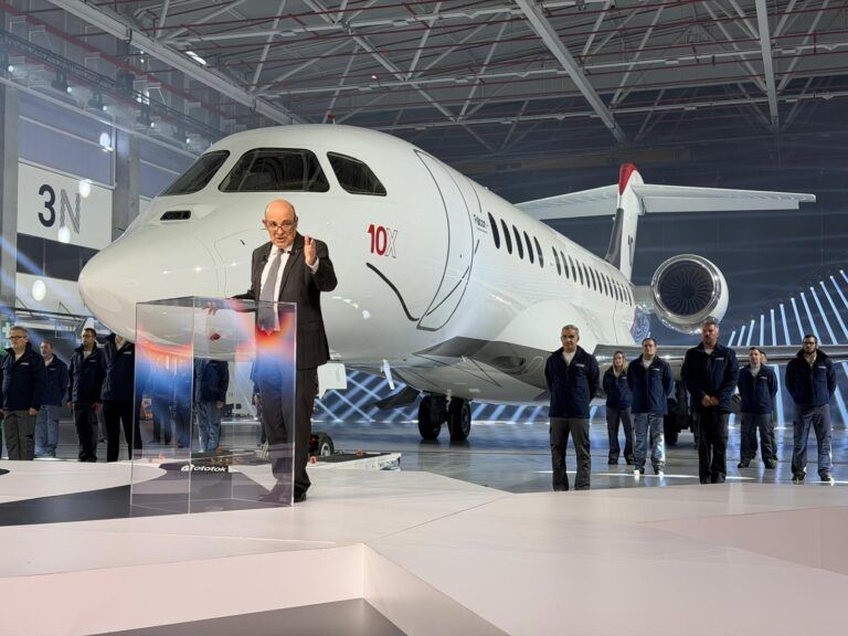 Eric Trappier speaks at a lectern in front of the Dassault Falcon 10X aircraft, which has just been unveiled in a large, brightly lit production hall, with workers standing alongside the aircraft in the background.