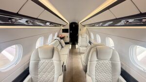 Interior view along the length of an Embraer Praetor 600 cabin showing cream leather seats, ambient LED lighting, and a three-seat divan at the far end.