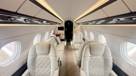 Interior view along the length of an Embraer Praetor 600 cabin showing cream leather seats, ambient LED lighting, and a three-seat divan at the far end.