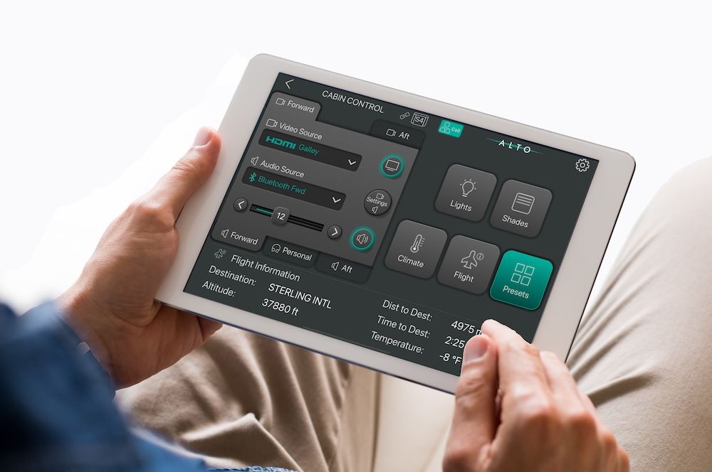 A passenger holds a tablet displaying the Alto Cadence system interface. The dark-themed interface shows controls for lights, shades, climate, flight information and entertainment, with teal accent colours. Flight data is visible at the bottom of the screen.