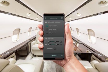 Alto Aviation reaches 100 aftermarket cabin audio installations on Falcon aircraft A hand holding a smartphone inside a business jet cabin with cream leather seats and wood-finish trim. The phone screen displays the Alto Aviation audio controls interface, showing MySound Optimisation settings with options for Full Cabin Tuned Stereo, VIP Focus Surround and VIP Row Surround, along with bass and treble tone controls and an Airshow Mode toggle.