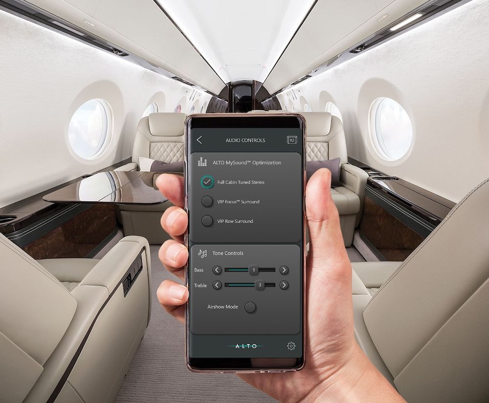 A hand holds a smartphone displaying the Alto MySound Optimisation interface, set against the interior of a private jet cabin. The app shows options including Full Cabin Tuned Stereo, VIP Focus Surround and VIP Row Surround, alongside bass and treble tone controls.