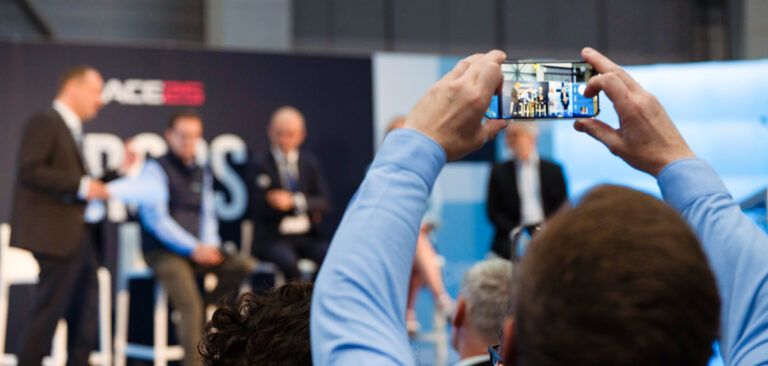 An audience member in a light blue shirt holds up a smartphone to photograph a panel discussion at the EBACE trade event, with speakers seated on stage in the background against branded signage