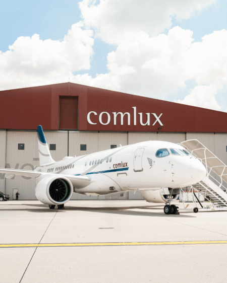 A white Airbus ACJ TwoTwenty aircraft in Comlux livery, featuring teal and grey accent striping and the Comlux logo on the fuselage, parked on the apron in front of a large brown and cream hangar building bearing the Comlux name. Passenger boarding stairs are positioned at the forward door. The sky is partly cloudy.