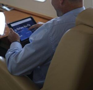 A male passenger seated in a business jet cabin uses a tablet device, with leather seating visible in the foreground
