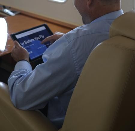 A male passenger seated in a business jet cabin uses a tablet device, with leather seating visible in the foreground