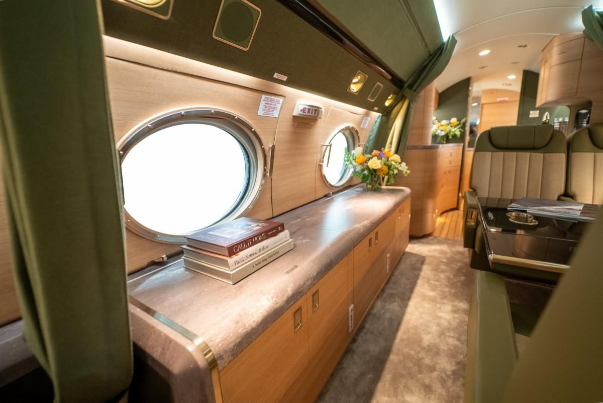 Interior view of a refurbished Gulfstream V cabin showing large circular porthole-style windows set into green fabric-lined sidewalls, a long marble-effect credenza with books and a vase of fresh flowers, light wood cabinetry, plush carpeting and leather seating with vertical channel stitching visible in the background.