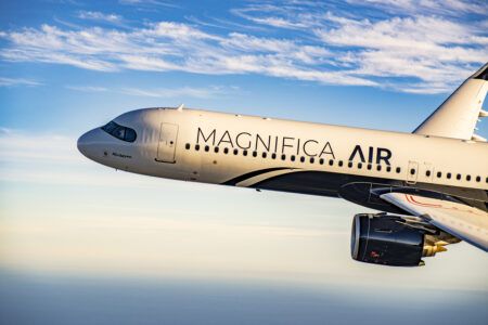 Close-up exterior view of a Magnifica Air ACJ321neo in flight against a blue sky with scattered clouds. The fuselage is finished in a light metallic livery with the Magnifica Air name in dark lettering. The forward fuselage, cockpit windows, passenger door and starboard engine are visible, with warm sunlight reflecting off the aircraft's surface.
