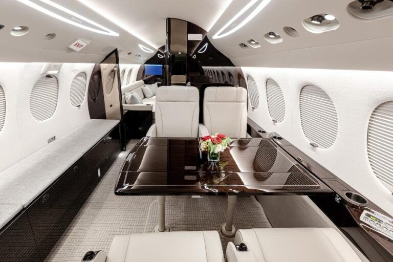 Duncan Aviation completes Falcon 7X interior refurbishment at Battle Creek facility Interior view of a refurbished Dassault Falcon 7X cabin looking aft, showing white leather seats arranged around a high-gloss dark wood conference table with a flower arrangement, light-coloured carpet with a subtle textured pattern, textured fabric upper sidewalls, oval cabin windows, and warm LED cabin lighting along the ceiling panels.