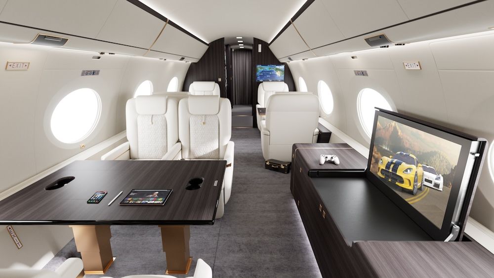 The interior of a large-cabin private jet, featuring white quilted leather seating, dark wood tables and a large flat-screen monitor displaying a racing scene, with a games console controller resting beside it. A tablet and smartphone are placed on the forward table, and a bulkhead-mounted screen is visible towards the rear of the cabin.
