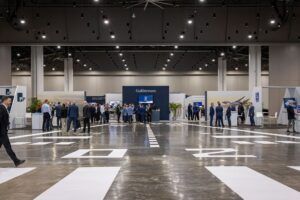 Wide-angle view of a large exhibition hall at the Savannah Convention Center during Gulfstream's Skyward event. Groups of attendees in business attire gather around exhibition stands, with a prominent navy blue Gulfstream-branded booth at the centre. Stands from Gogo and Rolls-Royce are visible to the left and right respectively. The polished concrete floor features white runway-style markings and the high ceiling is lit with rows of spotlights.