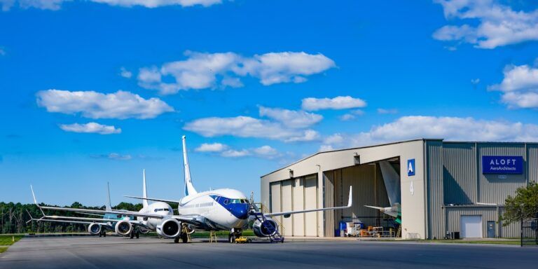 Two aircraft parked outside the Aloft AeroArchitects hangar on a sunny day, with the company name and logo visible on the building.